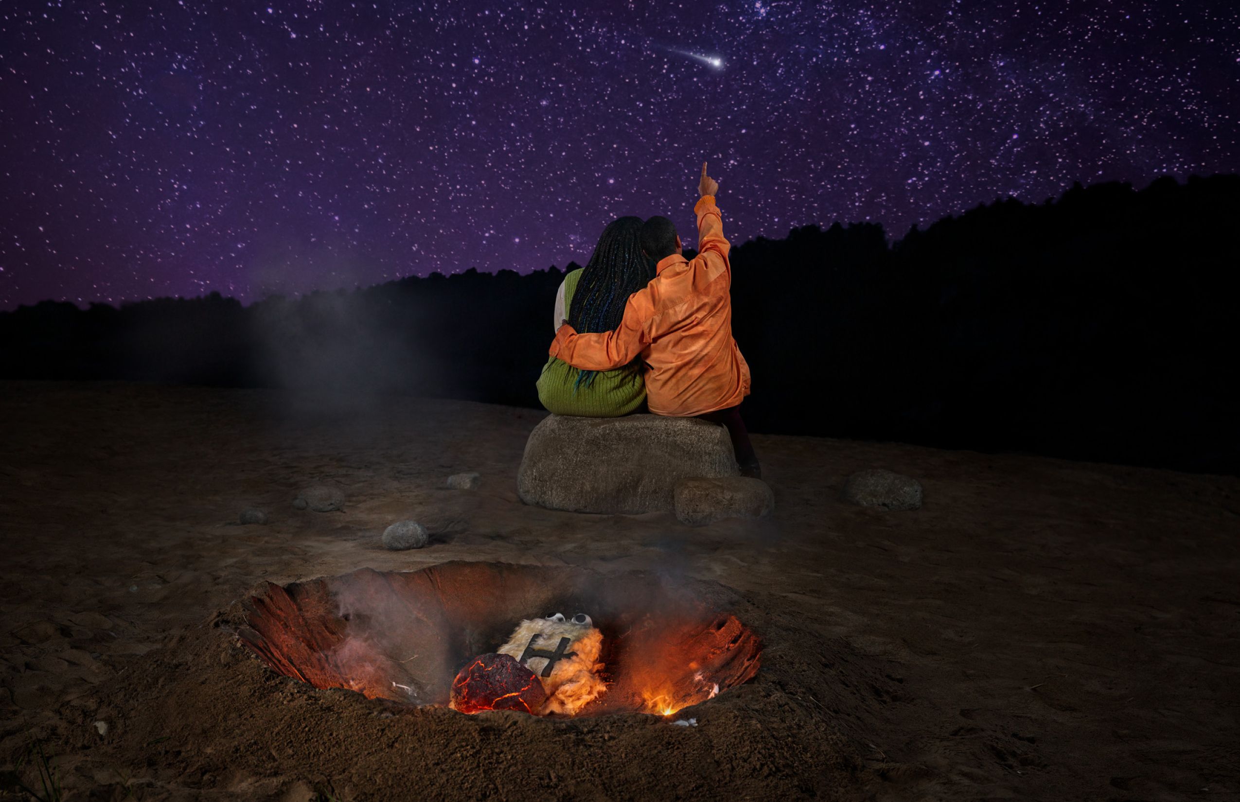 A couple staring up at the stars in the night sky while Hingie burns in a campfire