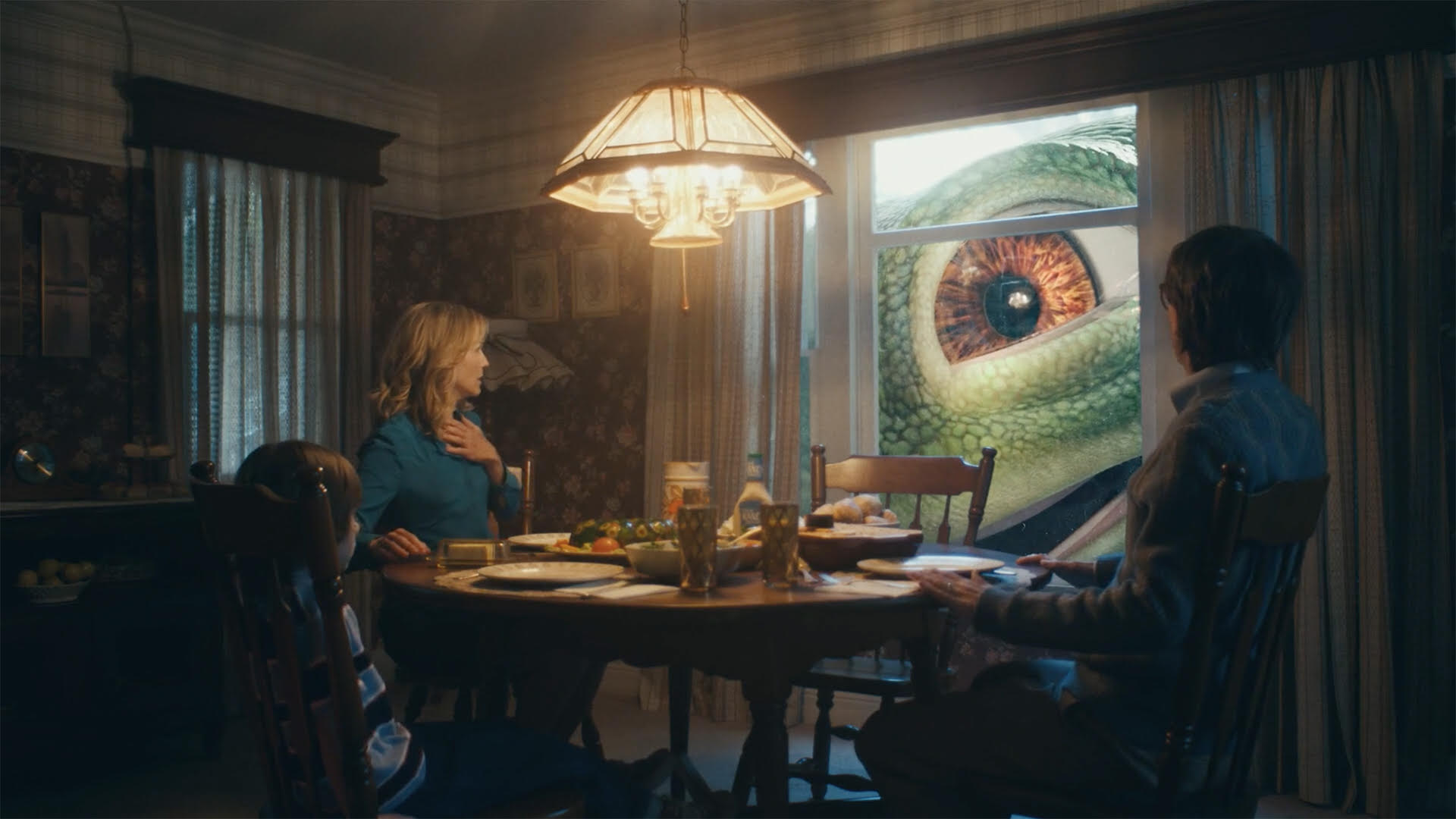 Parody of the Geico Gecko reimagined as dinosaur, looking in on couple having dinner